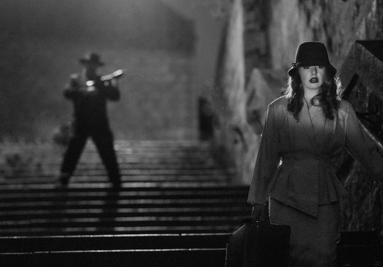 Escena de película en blanco y negro con una mujer en primer plano y un hombre detrás apuntándola con un arma.